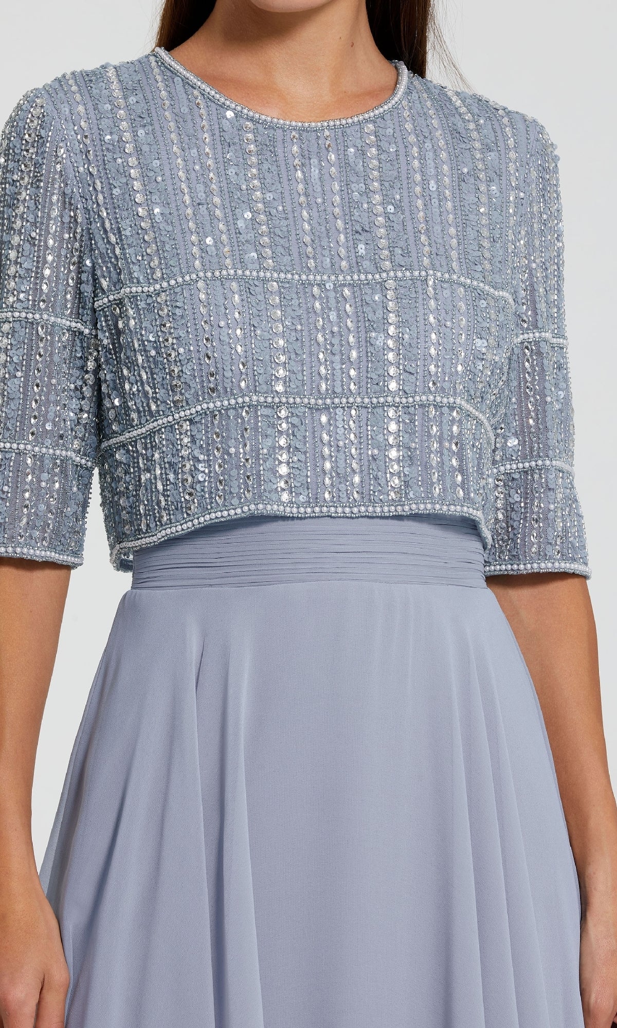 A person wearing the Mac Duggal 9180, a light blue long A-line formal dress with short sleeves and a beaded crop top featuring intricate vertical and horizontal sequin patterns.